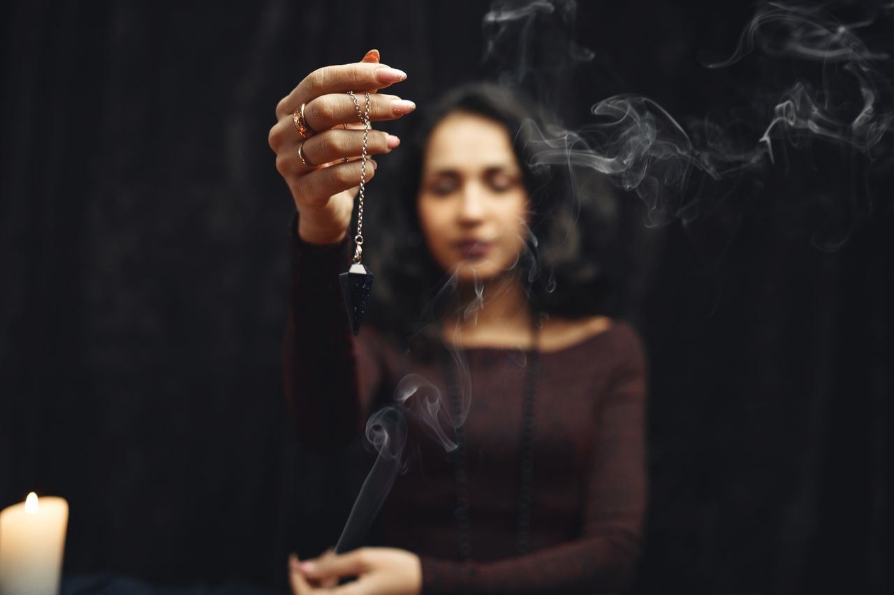 services-03 Close-up of a woman holding a pendulum over smoke, creating a mystical ambiance.